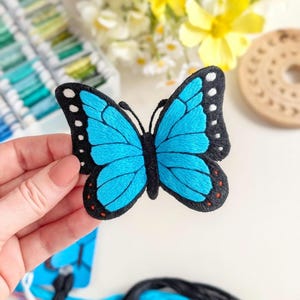 May include: An embroidered butterfly patch featuring vibrant blue wings with black borders, white spots along the edges, and small orange accents at the bottom. The butterfly has a black central body and two thin black antennae extending from the top.
