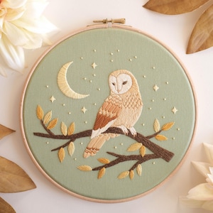May include: Embroidered owl art in a wooden hoop. The owl is perched on a branch with leaves, set against a light green background with a crescent moon and stars. The owl features brown and tan colours.