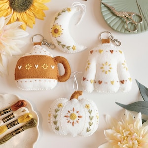 May include: A collection of handmade felt ornaments and keychains in autumnal colors. Includes a coffee cup keychain, a crescent moon, a sweater, and a pumpkin, all adorned with embroidered details. A plate of embroidery floss and a pair of scissors are also visible.