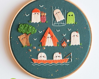 May include: Embroidered hoop art featuring a camping scene with ghosts, a tent, campfire, and boat. The design includes a tree, a jar, and a ghost in a cage. The background is teal, and the hoop is wooden. The text at the bottom says "Featuring glow-in-the-dark details."