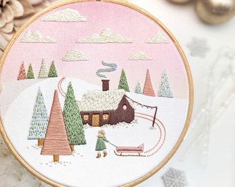 Peut inclure: Art de broderie en forme de cercle représentant une scène d'hiver avec une petite maison brune, des arbres enneigés et une personne tirant un traîneau. L'arrière-plan est un dégradé rose doux avec des nuages blancs.