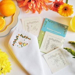 May include: A white linen napkin with embroidered orange and green cocktail design. The image also features a "Summer Corners Stick & Stitch" kit with floral and boat designs, along with orange and yellow flowers and a sliced orange.