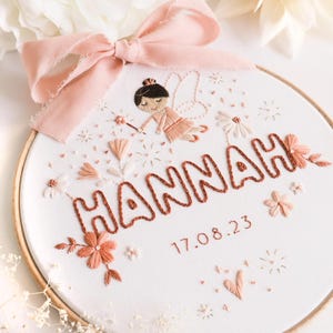 Può includere: Arte di ricamo su telaio con una fata con una bacchetta magica, fiori e il nome "HANNAH" in corsivo con la data "17.08.23" sotto.