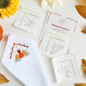 Könnte beinhalten: Eine weiße Stoffserviette mit einer Stickerei aus drei Kürbissen und Herbstblättern sowie eine Packung Tales from the Hoop Autumn Corners Stick- und Nähaufnäher mit Strichzeichnungen von Eicheln, Pilzen und Büchern.