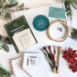 May include: A Christmas embroidery kit containing a green and white box, a wooden embroidery hoop, white fabric, embroidery floss in green, red, white, and gold, and a set of stick and stitch embroidery patches with Christmas corner designs. The box has the text "Tales from the Hoop" on it.