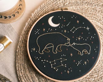 May include: Embroidered hoop art featuring two bears, a mother and cub, facing each other under a crescent moon and a starry night sky. The design is stitched in white thread on black fabric.