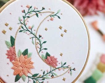 May include: A white embroidery hoop with a crescent moon design embroidered in gold thread. The moon is surrounded by green leaves and pink flowers. There are small gold stars scattered around the moon.
