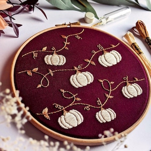 Könnte beinhalten: Stickrahmen-Kunst mit Stickerei, die einen burgunderfarbenen Stoff mit weißen Kürbissen und goldenen Ranken zeigt. Die Kürbisse sind in einem Kranz-Muster angeordnet.