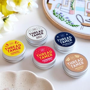 Puede incluir: Una colección de pequeñas latas redondas de metal con etiquetas coloridas. Cada lata está etiquetada como "Thread Tamer" con diferentes nombres de sabores como "Starburst", "Vanilla", "Strawberry", "Moonflower" y "Unscented". Las latas están dispuestas sobre una superficie blanca.