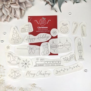 May include: A set of 14 white iron-on embroidery patches with Christmas themes. The patches feature designs such as a Christmas tree, a stocking, a train, a nutcracker, a candy cane, a bow, a snowflake, a bell, a rocking horse, a Christmas ornament, a wreath, and a string of lights. The patches are packaged on a red card with the text "Tales from the Hoop Christmas Stick & Stitch Embroidery Patches" and "Happy Holidays".