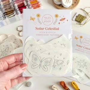 May include: A clear plastic package containing Solar Celestial stick and stitch embroidery patches with printed designs of butterflies, suns, and stars. The packaging features the text Tales From The Hoop and Solar Celestial Stick and Stitch Embroidery Patches.