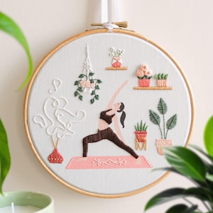 以下が含まれることがあります： ピンク色のマットの上でヨガのポーズをとる女性を描いた刺繍フープアート。デザインには鉢植えの植物、棚、抽象的な白い刺繍が施された花瓶が含まれています。このアートワークは木製のフープにセットされており、家の装飾に最適です。