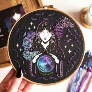 May include: An embroidered artwork in a wooden hoop, featuring a mystical theme. The design shows a robed figure with a crystal ball, with stars and swirling patterns in purple, blue, and gold. The background is black, creating a striking contrast.
