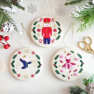 Op de afbeelding: Drie ronde, geborduurde kerstversieringen met gouden ophangdraden. Eén toont een rood-blauwe notenkraker, een andere een muis en de laatste een ballerina. Elk is omcirkeld door een groene krans met rode en roze details.