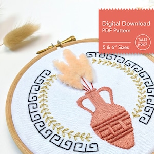 Könnte beinhalten: Stickerei auf einem Stickrahmen mit einem terracottafarbenen Vasenmotiv mit einem stilisierten Design, umgeben von einem goldenen und schwarzen griechischen Schlüsselmuster. Die Vase ist mit einer flauschigen, pfirsichfarbenen Blume gefüllt. Der Rahmen ist aus Holz und hat einen weißen Stoffhintergrund. Der Text "Digitaler Download PDF-Muster" und "5 & 6" Größen" ist in der oberen rechten Ecke sichtbar.