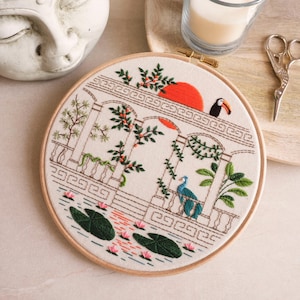 Könnte beinhalten: Stickerei in einem runden Rahmen mit einer detaillierten Szene eines weißen Gebäudes mit einer roten Sonne, grünen Pflanzen, einem blauen Pfau und einem Tukan. Die Szene ist von einem braunen Holzrahmen umgeben.