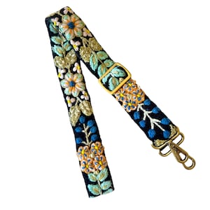 May include: A black fabric bag strap featuring intricate floral embroidery in shades of orange, light blue, and green. The strap includes an adjustable gold-toned metal slider and two brass-colored swivel lobster clasps at one end for attachment.