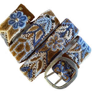 May include: A decorative belt with a floral design. The belt features a brown base with blue and white embroidered floral patterns. The buckle is silver-colored. The belt is approximately 1.5 inches wide.