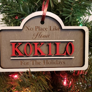 May include: A wooden Christmas ornament with a white border and a red ribbon. The ornament is shaped like a sign and says "No Place Like Home" with the name "KOK1LO" in red letters and "For The Holidays" below.