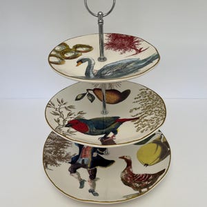 May include: A three-tiered ceramic cake stand with a silver metal handle. Each tier features a different whimsical design with birds, fruit, and other nature-inspired elements.