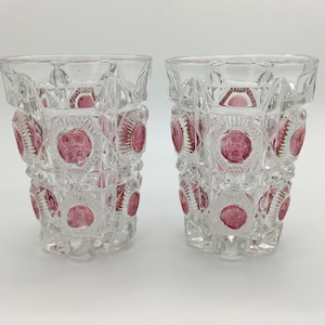 May include: Two clear, cut-glass tumblers with a repeating pattern of pink circles and geometric shapes. The glasses have a thick base and a straight, slightly flared rim. The design is symmetrical and the pink colour adds a pop of colour.