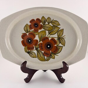 May include: Oval serving platter with a floral design. The platter features a beige speckled background with a pattern of orange flowers and green and yellow leaves. The platter is displayed on a dark brown wooden stand.