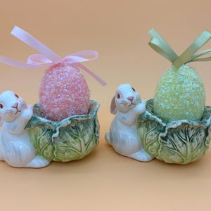May include: Two ceramic Easter bunny ornaments, each holding a leaf-shaped bowl with a decorative egg. One egg is pink with a pink ribbon, the other is green with a green ribbon. The bunnies are white with pink details.
