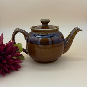 Peut inclure: Une théière en céramique marron avec un couvercle, une anse et un bec verseur. La théière a une finition brillante et une bande marron plus foncée au milieu. Une fleur bordeaux avec des feuilles vertes est en arrière-plan. La théière est conçue pour infuser du thé.