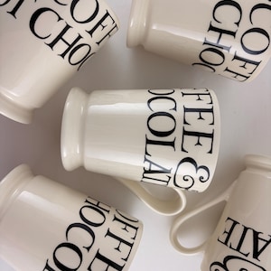 May include: Cream ceramic mugs with black lettering spelling out "COFFEE", "TEA", and "HOT CHOCOLATE". The mugs have a traditional shape, a handle, and a gently curved rim. The mugs are arranged on a white surface.