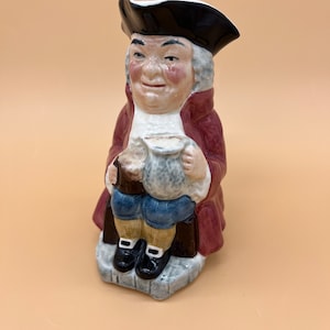 May include: A ceramic figurine of a man wearing a red coat and a black hat, holding a mug in each hand. The figurine is sitting on a chair and has a detailed facial expression.