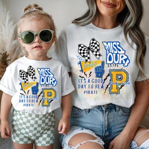 May include: White t-shirts with a graphic design. The design includes the text "MISS OUR", "65186", "It's a good day to be a Pirate", a checkered bow, and a blue and yellow "P" logo. The shirts are worn by a child and an adult.