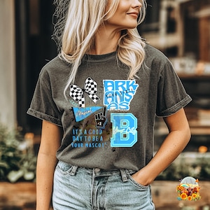 May include: Dark grey t-shirt with a graphic design featuring the text "ARK YOUR STATE" in blue, a checkered bow, and the text "IT'S A GOOD DAY TO BE A YOUR MASCOT". The shirt is worn by a person.