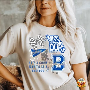 May include: Cream-colored t-shirt with a graphic design featuring a checkered bow, the word "Bulldogs", a raised foam finger, paw prints, and the words "It's a good day to be a Bulldog". The word "Missouri" is also present.