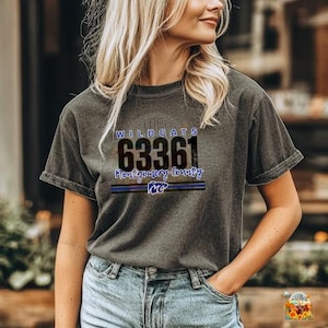 Könnte beinhalten: Dunkelgraues T-Shirt mit dem Text "WILDCATS 63361 Montgomery County MO" in Blau und Schwarz. Das Shirt ist ein kurzärmeliger Rundhalsausschnitt.