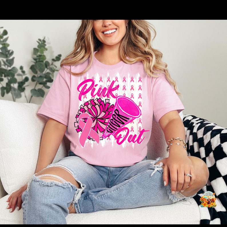 以下が含まれることがあります： ピンク色のTシャツ。ピンクのリボン、ポンポン、メガホンが描かれたグラフィックデザイン。Tシャツのテキストは「Pink Cheer Out」と書かれています。