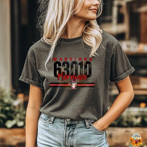 Puede incluir: Camiseta gris oscuro con la palabra "WARRIORS" encima del número "63010" y la palabra "Meramec" en rojo. Una franja roja y negra con un gráfico de tejón está debajo del texto. La camiseta es de material suave.