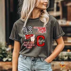 May include: Dark grey t-shirt with a graphic design featuring the text "OKLAHOMA", "Comets", and "Hurton". The design includes a checkered bow, a raised index finger, and the phrase "IT'S A GOOD DAY TO BE A COMET".