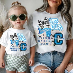 Peut inclure: Deux t-shirts blancs avec des graphiques assortis. Les chemises présentent un nœud drapeau à damier, le texte "MISS OUR", "IT'S A GOOD DAY TO BE A COUGAR" et un "C" bleu avec des empreintes de pattes. Un t-shirt est porté par un enfant.