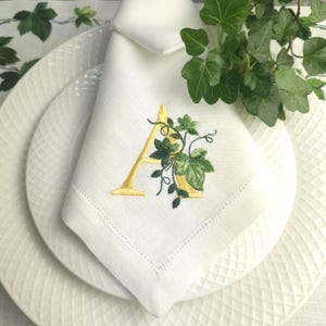 Pode incluir: Guardanapo de linho branco com a letra "A" bordada em amarelo e um desenho de hera verde. O guardanapo é dobrado e colocado sobre um prato branco com um padrão texturizado. Ramos de hera fresca ao fundo.