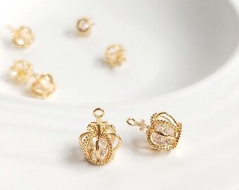 以下が含まれることがあります： 透明なクリスタルアクセントが付いた、2つの金色のクラウン型のチャームのクローズアップ。チャームは白い表面にあります。