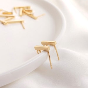 May include: Two gold bar earrings with a simple design. The earrings are on a white surface.