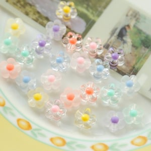May include: A collection of pastel-coloured plastic flower charms. The charms are translucent and have a 3D flower shape. The flowers are in various colours, including pink, blue, yellow, green, and purple.