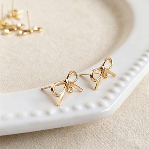 May include: A pair of gold-coloured bow-shaped stud earrings. The earrings are displayed on a white, scalloped-edge plate. Additional earring components are visible in the background. The earrings have a post and loop design.