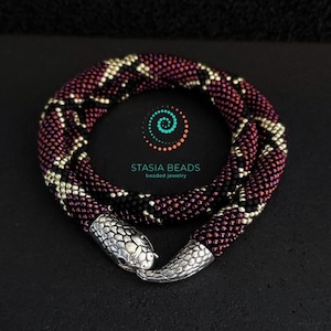Peut inclure: Un bracelet de perles bordeaux, noir et argenté, avec un fermoir en forme de tête de serpent. Le bracelet est tissé de manière complexe avec de petites perles, affichant un motif. Le logo "STASIA BEADS beaded jewelry" est visible.