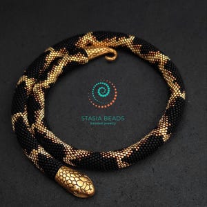 Puede incluir: Un collar flexible con forma de serpiente hecho de pequeñas cuentas negras y doradas metálicas, con una cabeza de metal en tono dorado texturizado y una cola curva. La pieza muestra el texto STASIA BEADS beaded jewelry con un logotipo en espiral.