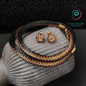 Könnte beinhalten: Ein Schmuckset mit Perlen, bestehend aus einem Armband und passenden Ohrringen. Das Armband hat ein Spiraldesign mit goldenen, schwarzen und schillernden Perlen. Die Ohrringe sind klein, rund und goldfarben. Der Schmuck wird auf einer strukturierten, grauen Oberfläche präsentiert.