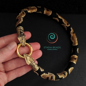 May include: A beaded necklace with a snake design. The necklace features a black and gold pattern, with gold snake head clasps and a gold ring clasp. The text "STASIA BEADS beaded jewelry" is visible.