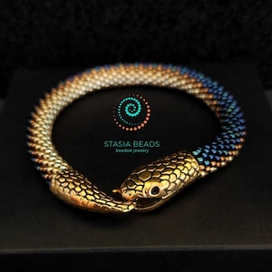 Op de afbeelding: Een gouden slangenarmband met een kralenlichaam in tinten blauw, wit en goud. De armband is gemaakt door Stasia Beads, een bedrijf voor kralenjuwelen.