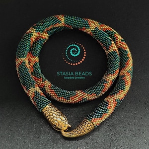 May include: A beaded snake necklace with a gold snake head clasp. The necklace is made with green, orange, and gold beads. The necklace is on a black background. The text "Stasia Beads Beaded Jewelry" is visible in the image.