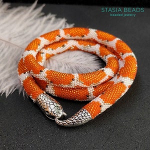 May include: Orange and white beaded snake necklace with a silver snake head clasp. The necklace is coiled on a black surface with a white feather in the background. The text "STASIA BEADS beaded jewellery" is visible in the top right corner of the image.